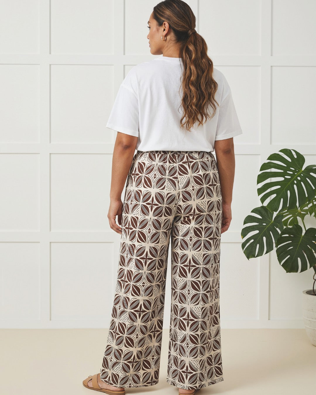 Woman wearing a white t-shirt and patterned pants standing in a room with a plant.