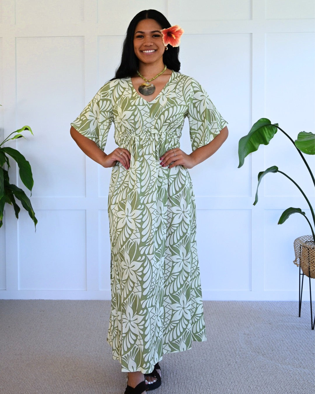 Woman wearing a green and white patterned island dress standing in a room with plants.