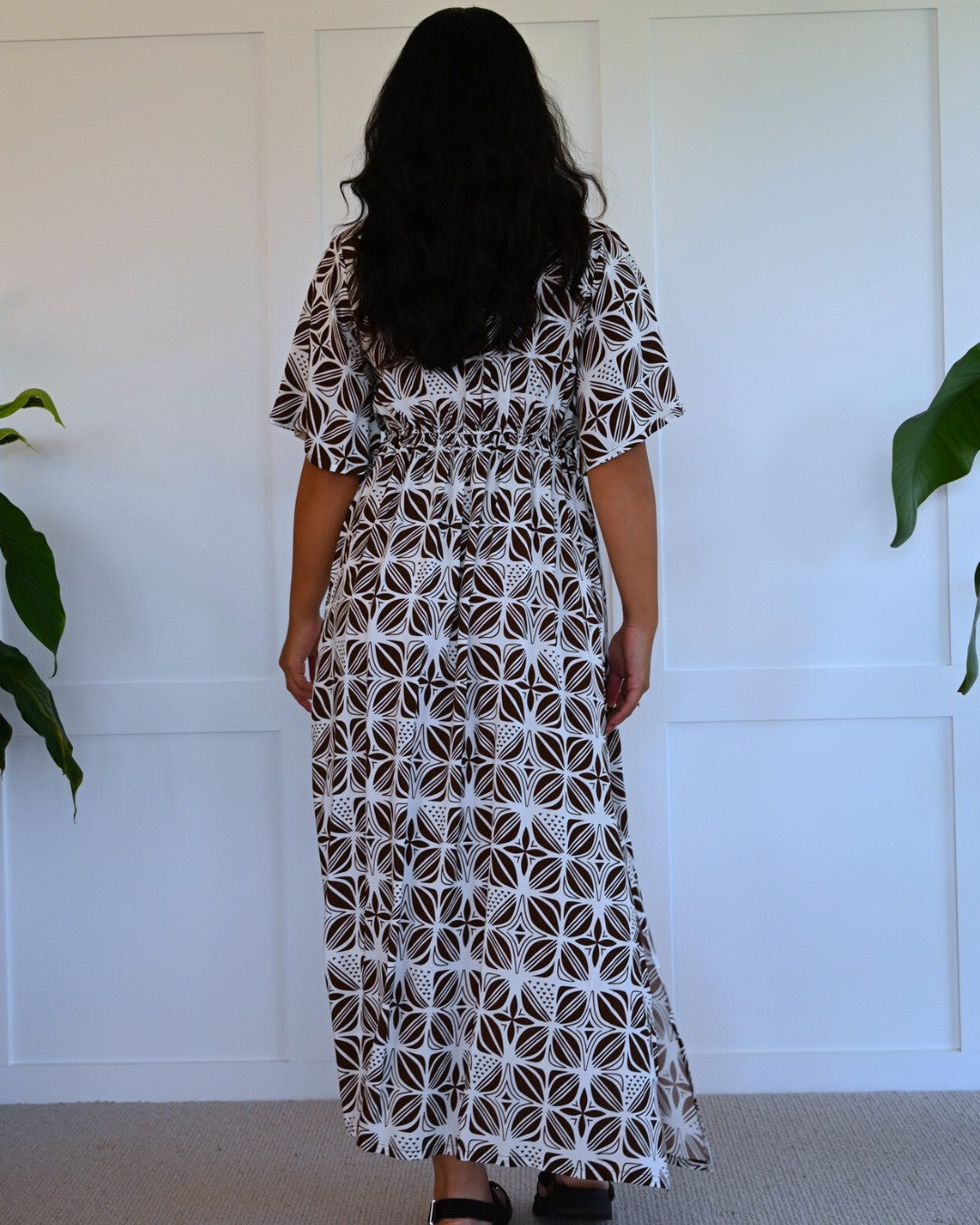 Woman wearing a patterned island dress standing against a white wall with plants.