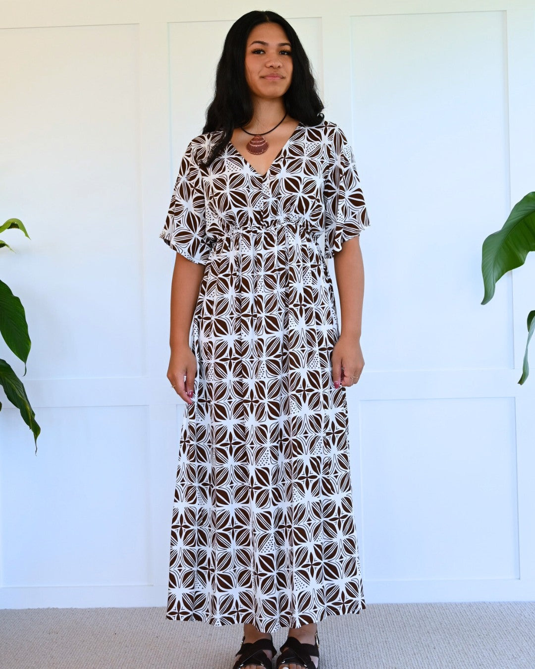 Woman wearing a patterned island dress standing against a white wall with plants.