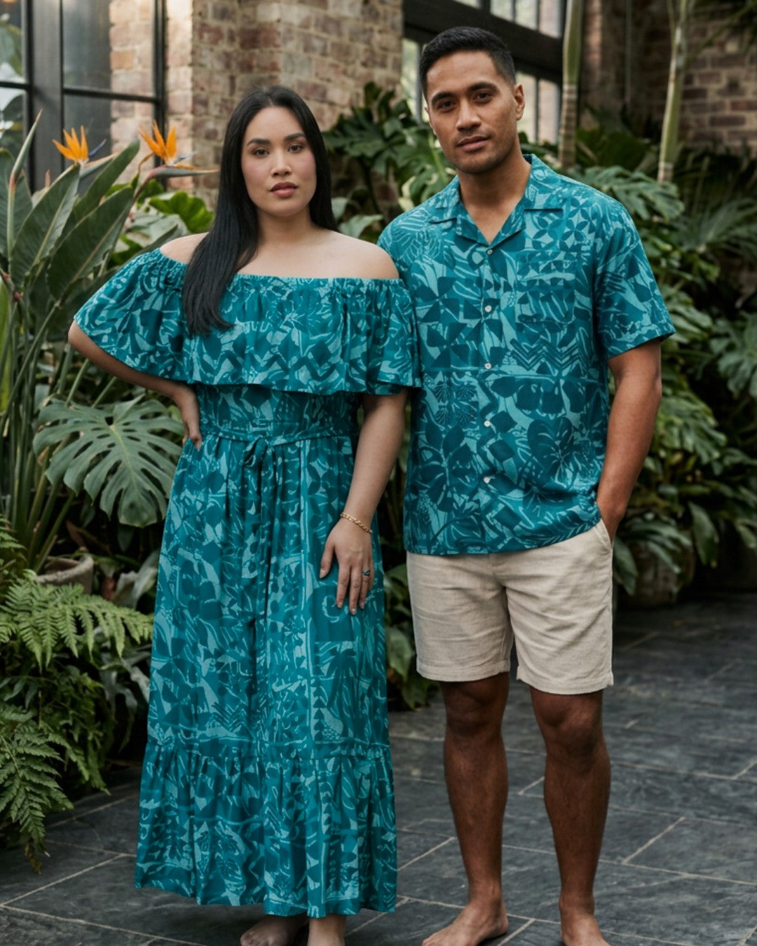 A man and a woman modeling matching teal tropical leaf print clothing in an outdoor garden setting. The woman wears an off-the-shoulder maxi dress, and the man wears a matching short-sleeve button-up shirt with beige shorts.