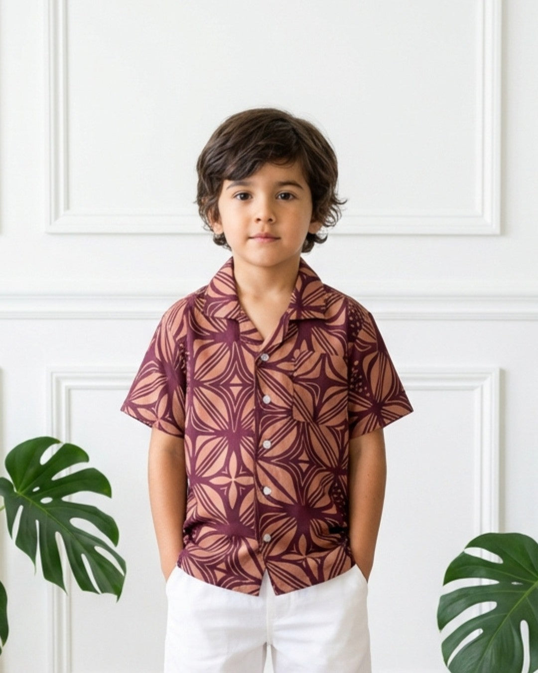 Child wearing a patterned shirt standing against a white wall with green leaves.