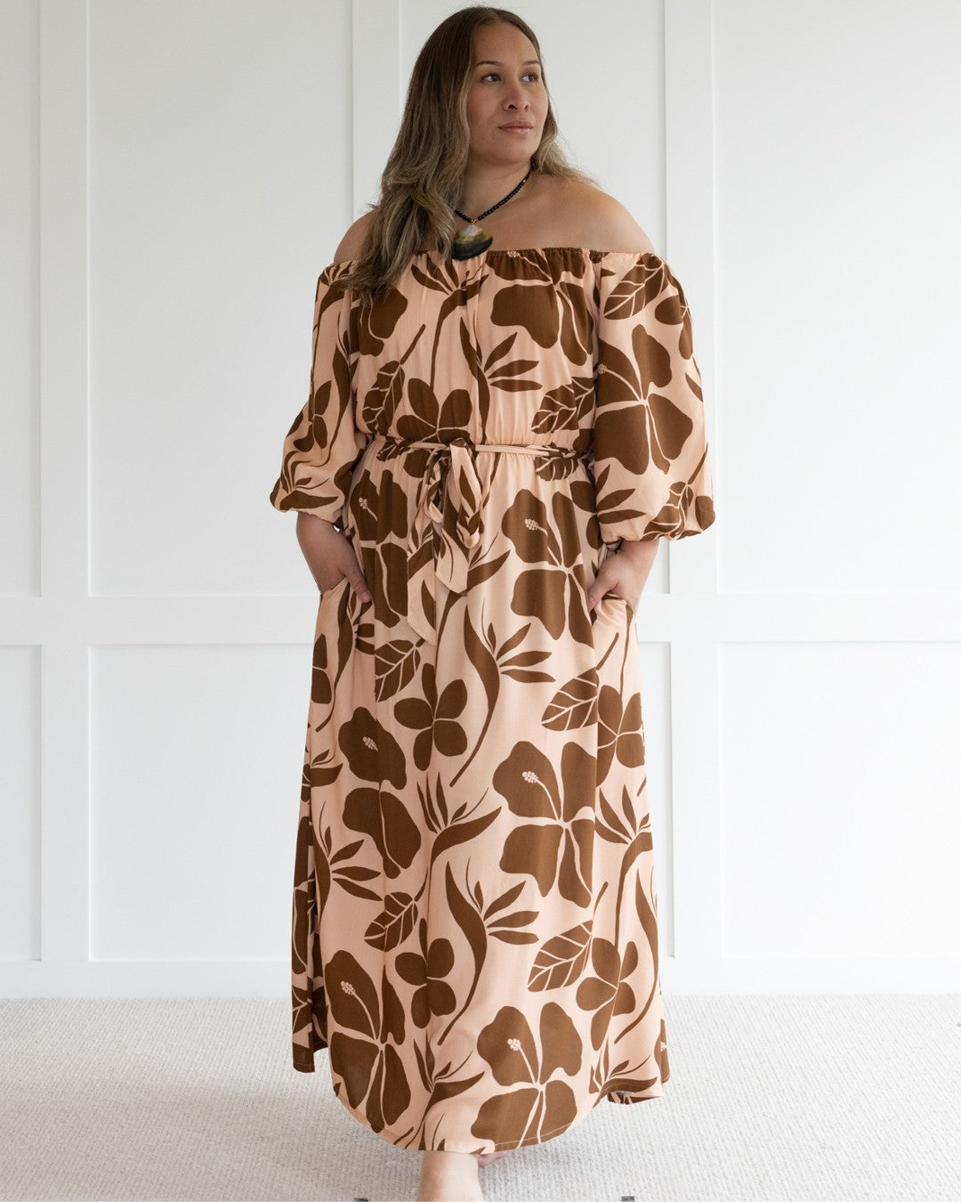 Woman wearing a floral off-shoulder island dress against a white wall.