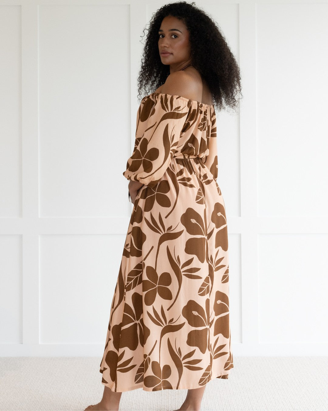 Woman wearing a floral off-shoulder island dress against a white wall.