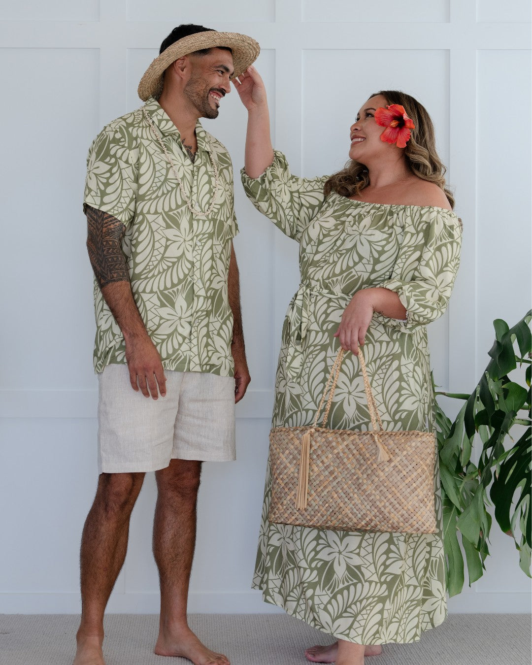 Man and woman wearing matching green leaf-patterned island wear standing against a white background.