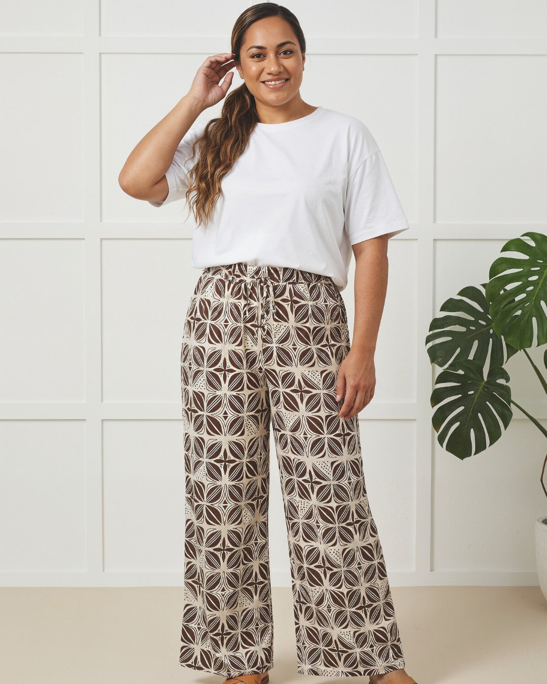 Woman wearing a white t-shirt and patterned pants standing in a room with a plant.