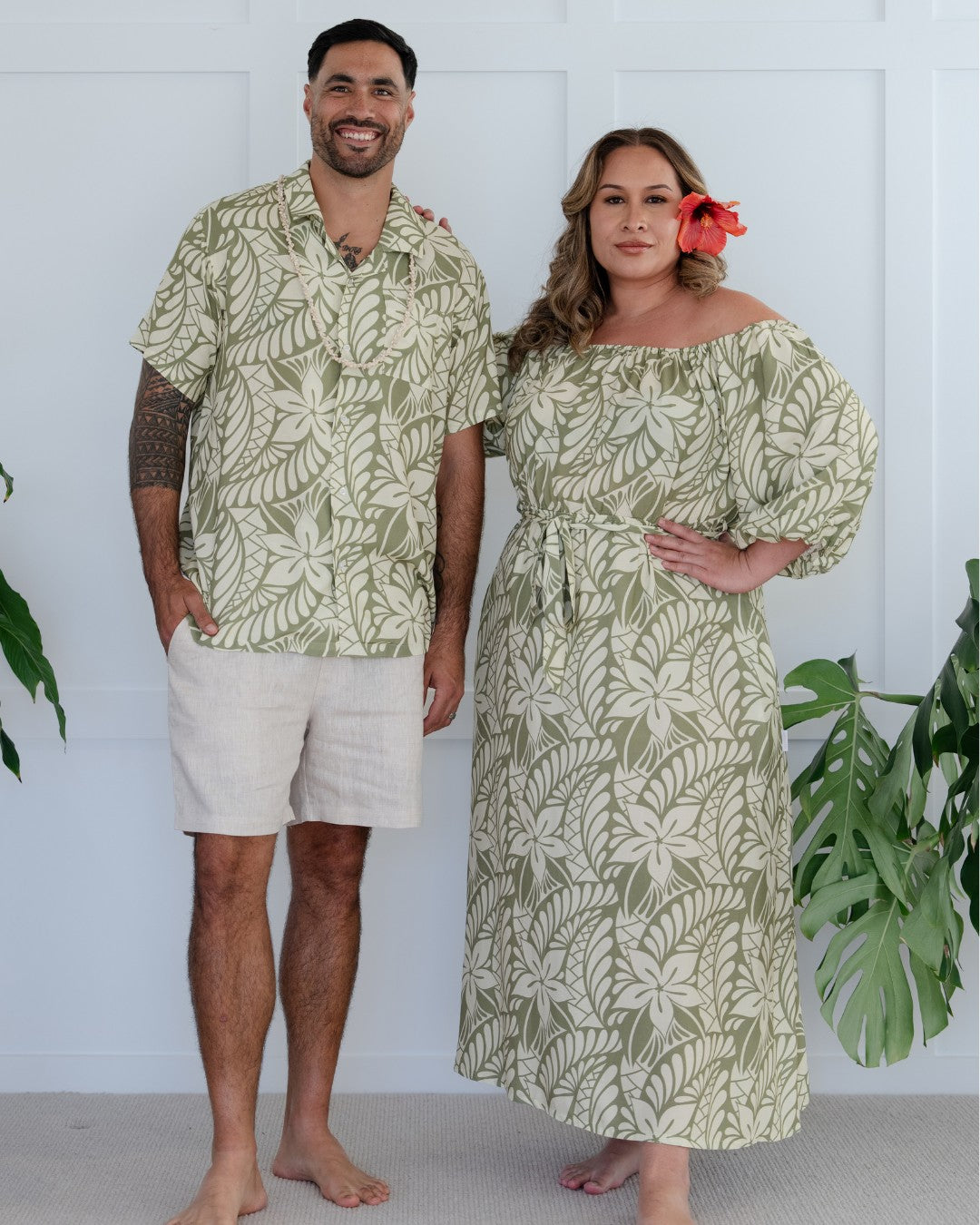 Man and woman wearing matching green leaf-patterned island wear outfits standing against a white background with plants.