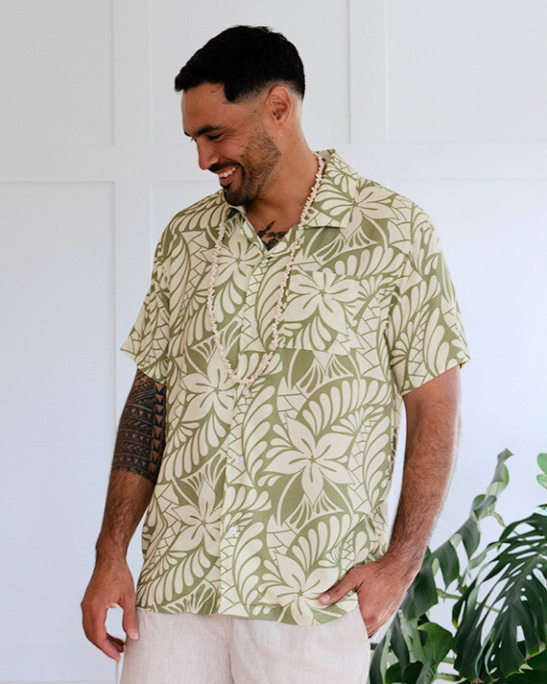 Man wearing a patterned island shirt standing against a white wall with plants in the background