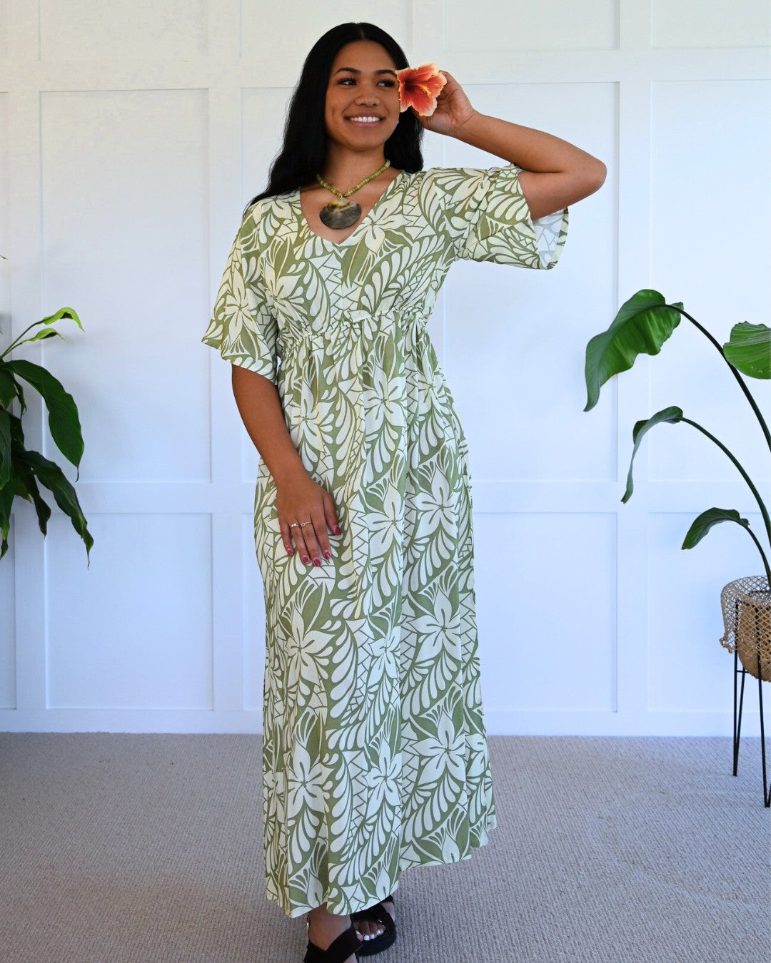 Woman wearing a green and white patterned island dress standing in a room with plants.