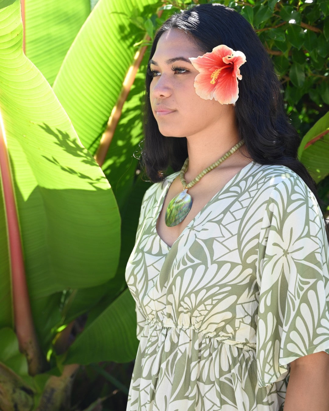 Woman wearing a island floral dress with a flower in her hair, standing among green foliage.