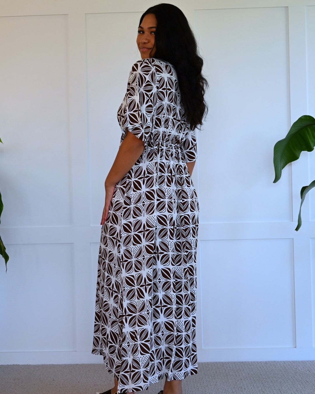 Woman wearing a patterned island dress standing against a white wall with plants.