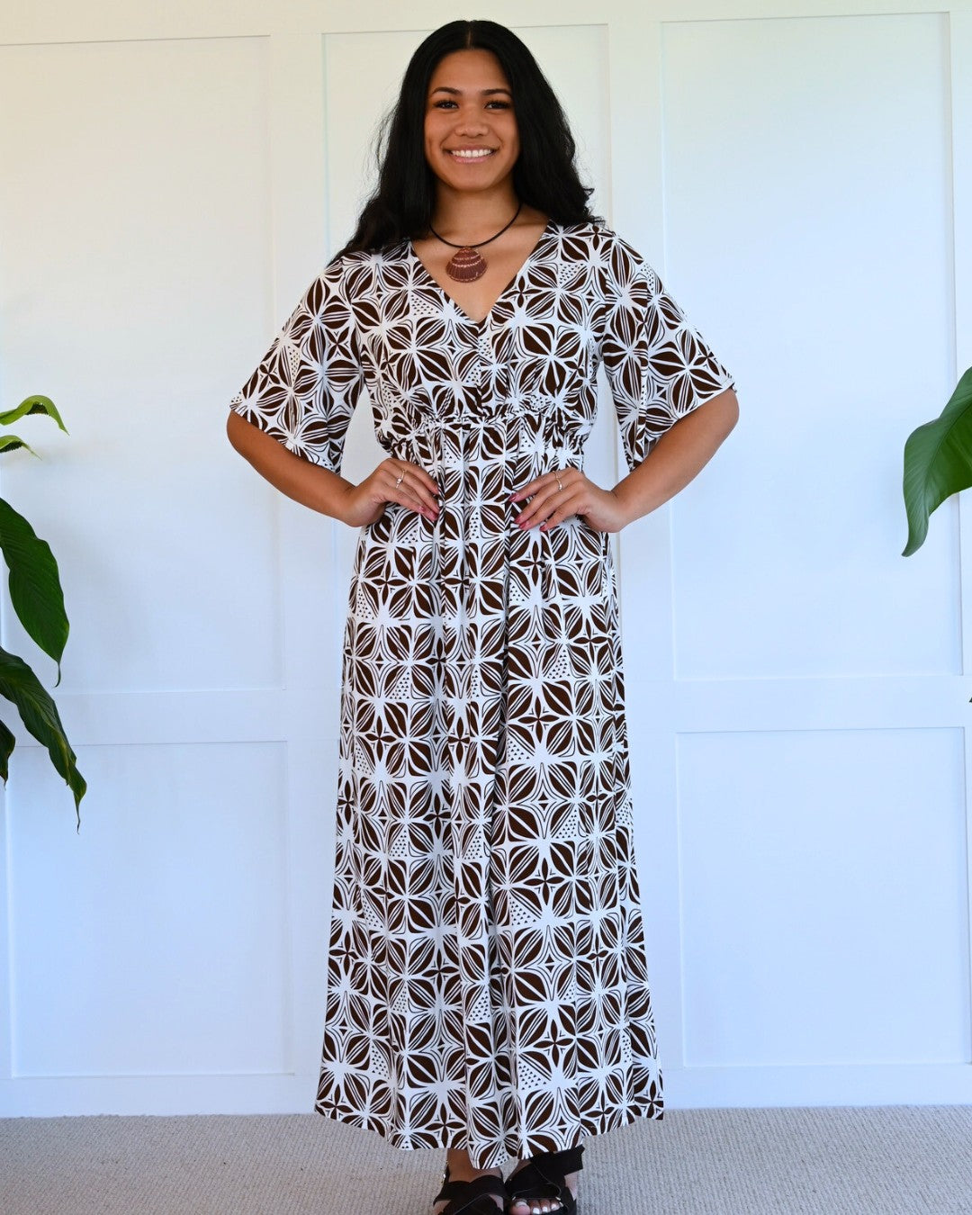 Woman wearing a patterned island dress standing against a white wall with plants.