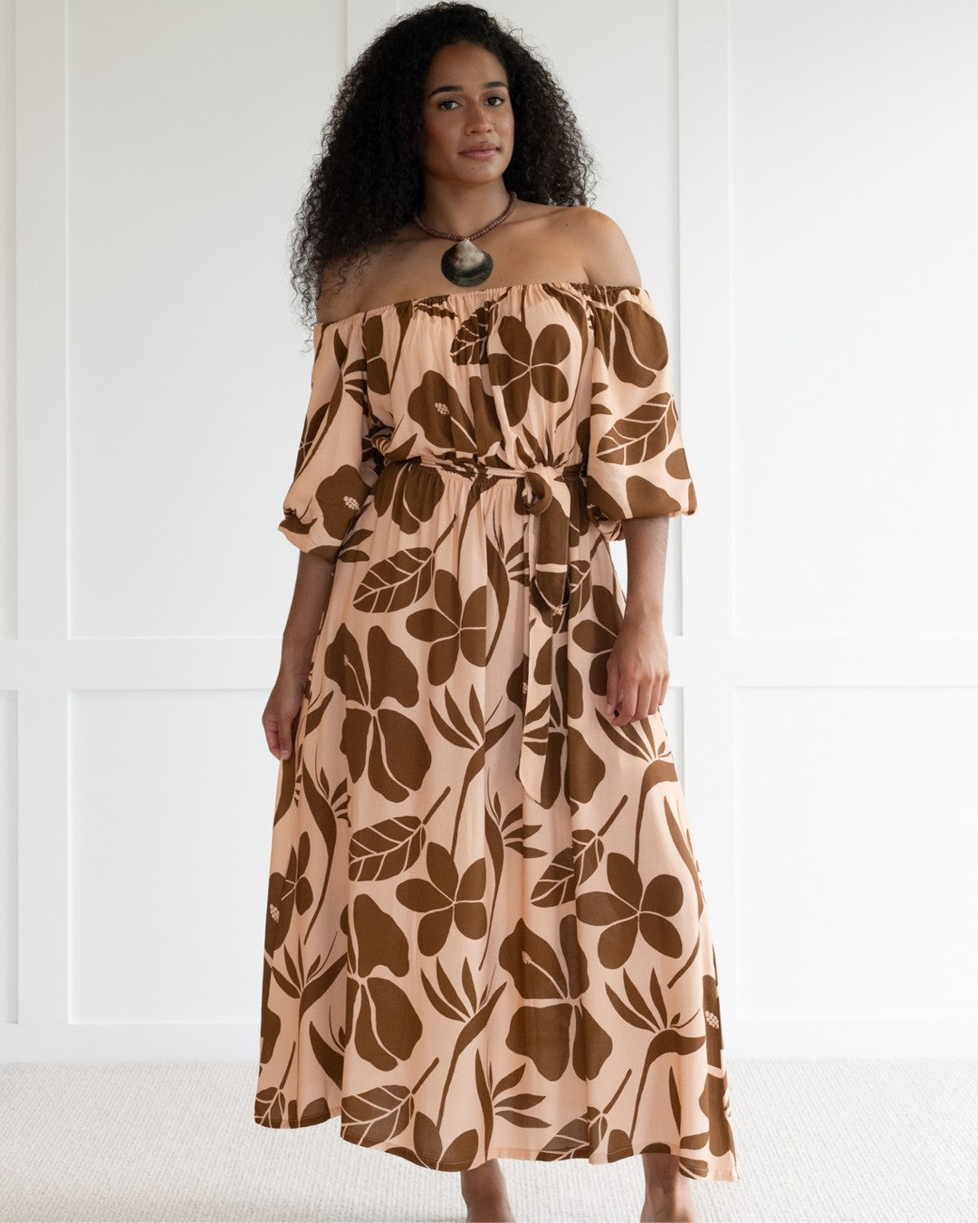 Woman wearing a floral off-shoulder island dress against a white wall.